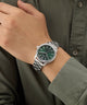 Hand wearing a silver GUESS Mens Silver Tone Analog Watch with a green face on a person wearing a green shirt.