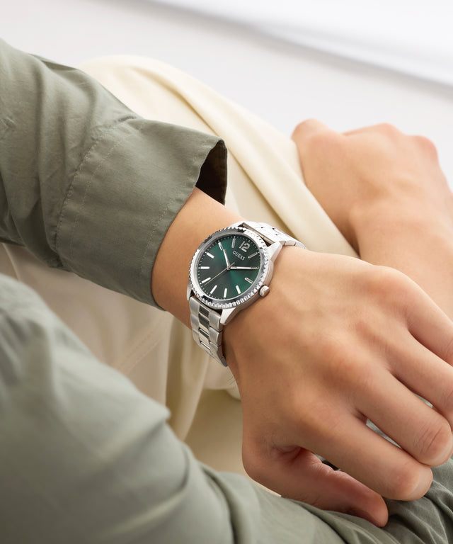 Person wearing a silver GUESS Mens Silver Tone Analog Watch with a green dial on a light background