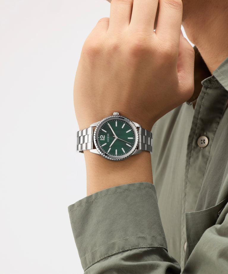 Hand wearing a GUESS Mens Silver Tone Analog Watch with a green face and silver band on a plain background