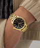 Gold GUESS Mens Gold Tone Analog Watch with a black face on a wrist, wearing a beige jacket.