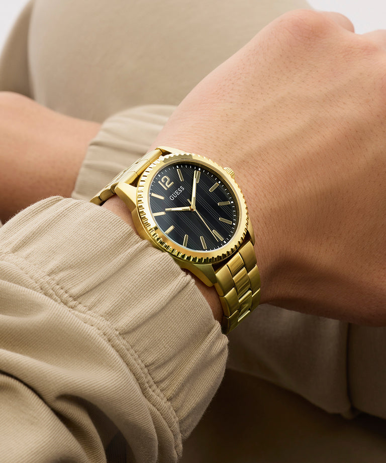 Gold GUESS Mens Gold Tone Analog Watch with black dial on a wrist, wearing a beige jacket.