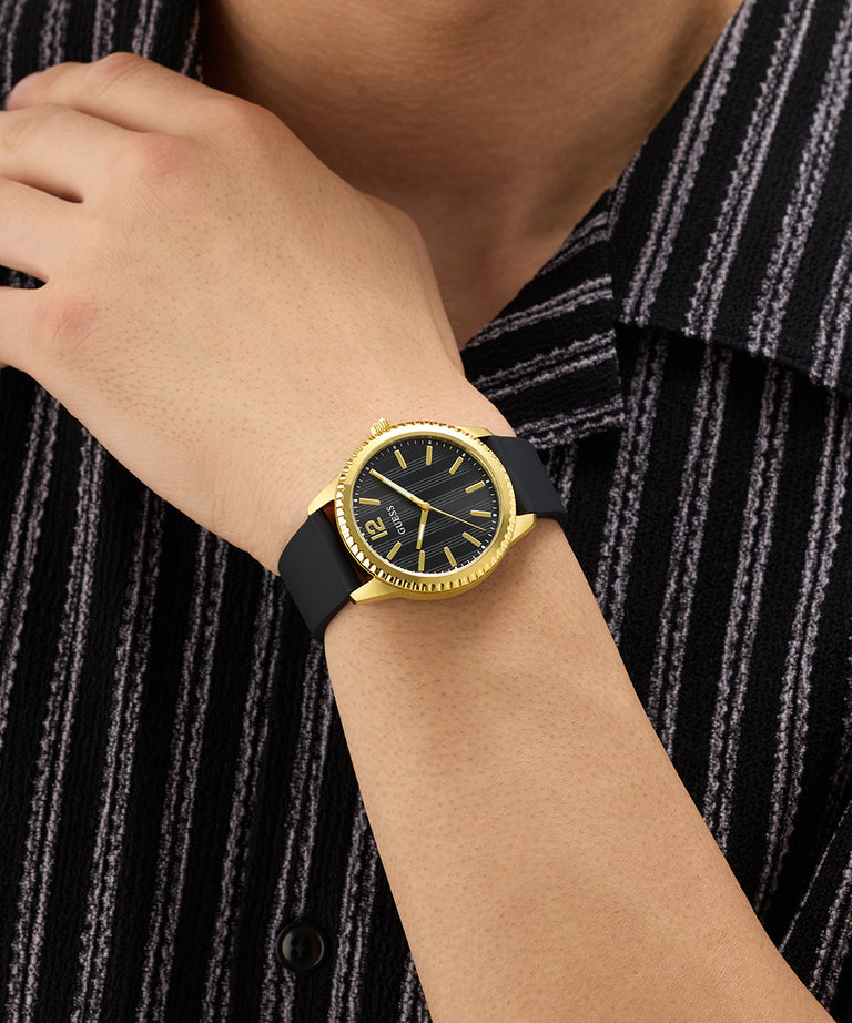 Gold GUESS Mens Black Gold Tone Analog Watch with black strap on a wrist against a striped shirt background