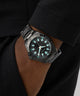 Black wristwatch with blue glowing face on a person's wrist against a dark background