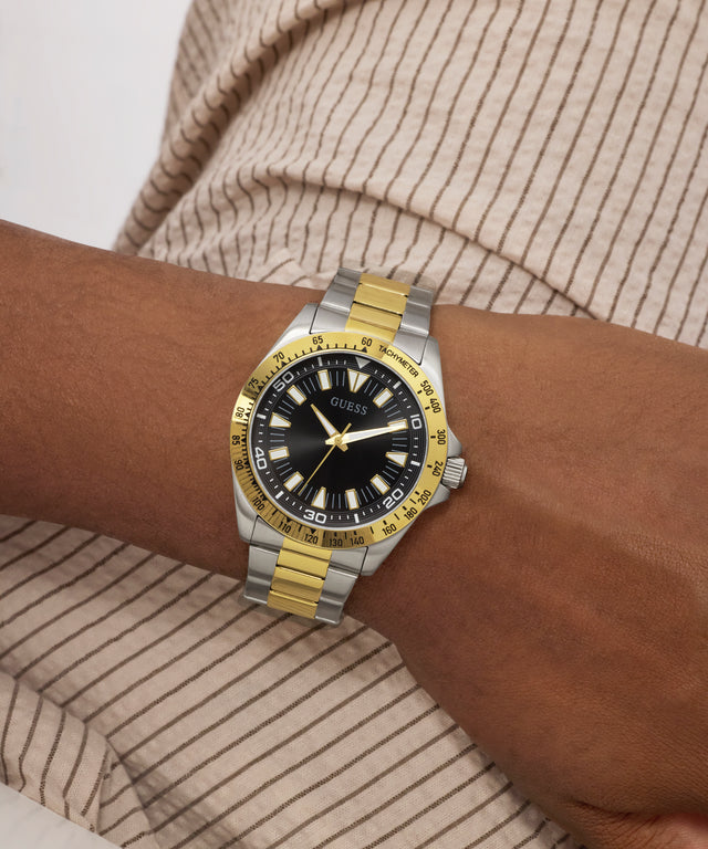 Wrist wearing a Guess watch with a gold and silver band on a neutral background