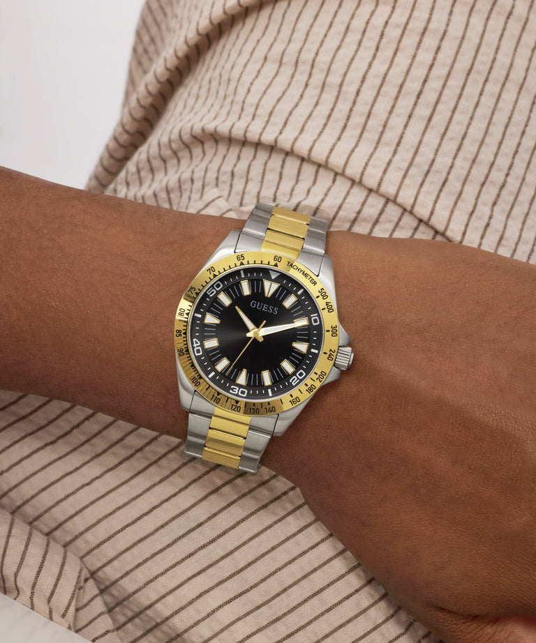 Wrist wearing a Guess watch with a gold and silver band on a neutral background