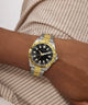 Wrist wearing a Guess watch with a gold and silver band on a neutral background