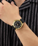 Gold watch with black strap on a wrist against a striped background