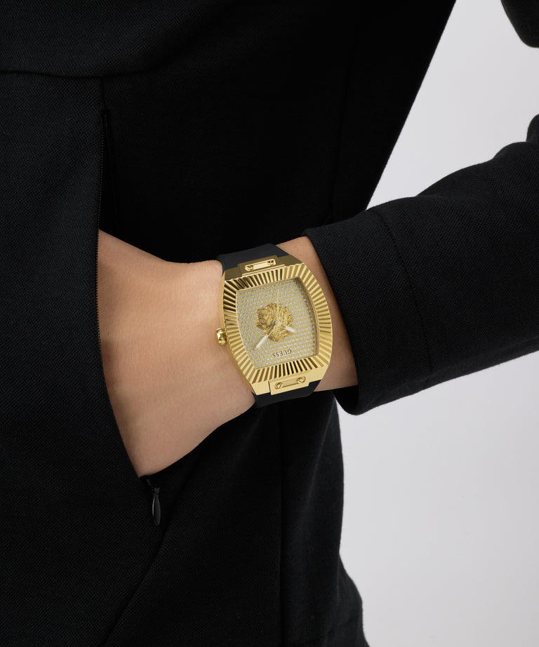 Gold GUESS Mens Black Gold Tone Analog Watch with a lion emblem on a person wearing a black outfit against a white background