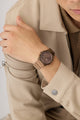 Person wearing a brown GUESS Mens Jaq Coffee Analog Watch on a beige jacket against a neutral background