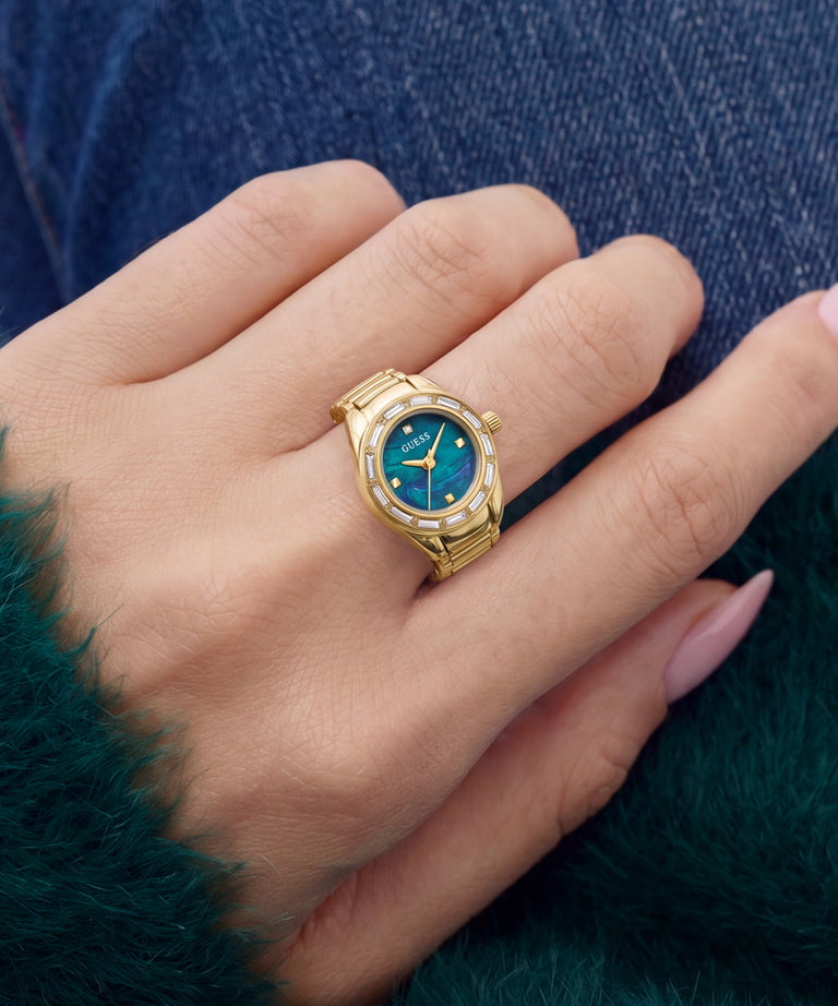 A gold-tone watch with a blue dial and bezel, worn on a person's finger. The watch has a gold mesh band and a bezel accented with small clear stones.