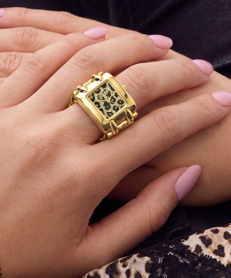 A close-up of a hand wearing a chunky gold ring with a square face and black leopard print detailing, resting on another hand with pink manicured nails.