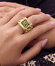 A close-up of a hand wearing a chunky gold ring with a square face and black leopard print detailing, resting on another hand with pink manicured nails.