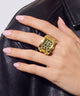 A close-up of a hand with pale pink manicured nails wearing a large gold ring with a rectangular face featuring a black leopard print pattern, resting on a black leather surface.