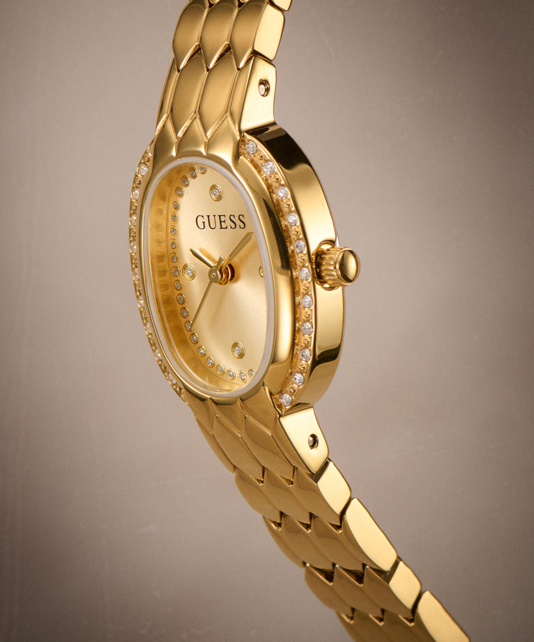 Gold-tone Guess watch with a diamond-accented bezel and matching gold-tone link bracelet, shown at an angle against a neutral gray background.