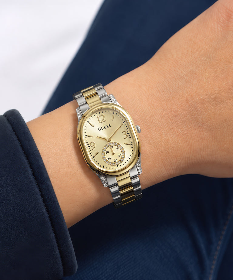 A gold and silver two-tone Guess watch with a gold dial and bezel, worn on a person's wrist against a navy blue and white background.