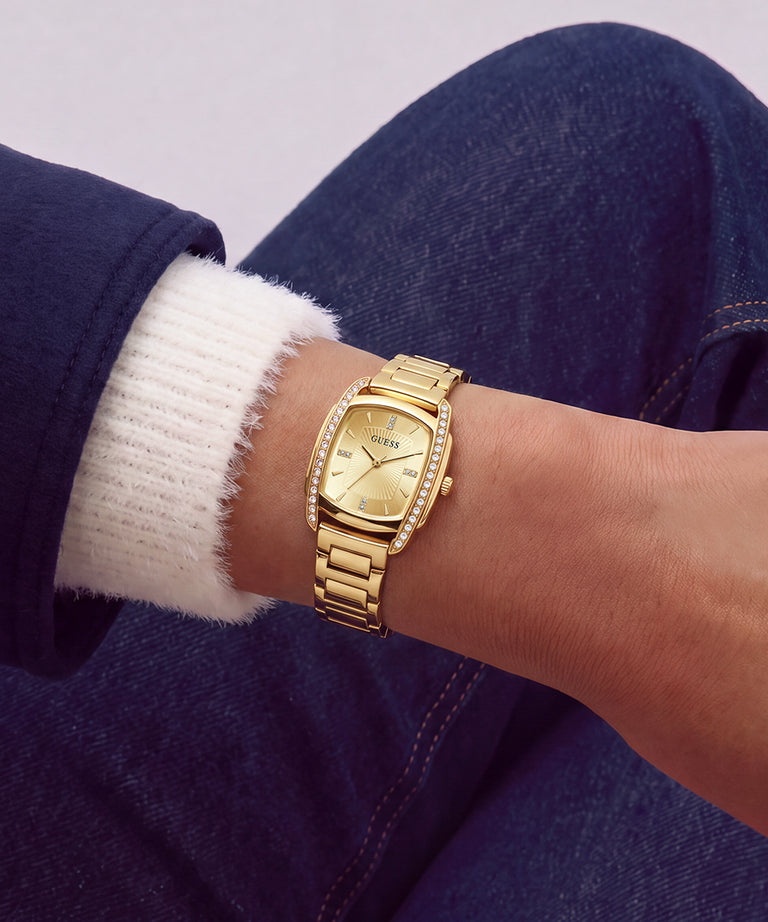 A gold-tone GUESS watch with a rhinestone-encrusted bezel and matching metal link bracelet, worn on a person's wrist. The person is wearing a white ribbed knit sweater cuff and dark blue denim jeans.