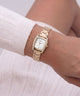 A gold-tone women's watch with a white dial and crystal accents, shown on a model's wrist wearing a white ribbed knit sweater.
