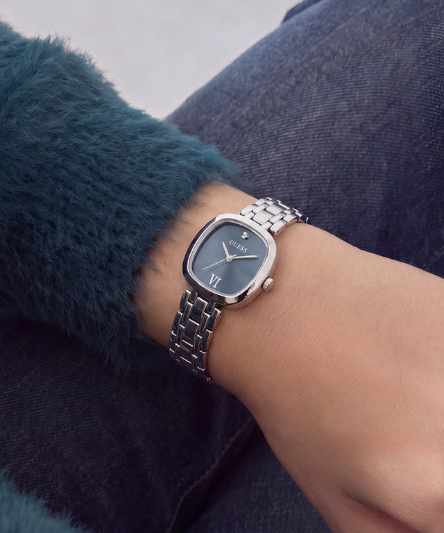 A silver-tone metal link watch with a dark blue dial and Roman numeral markers, worn on a person's wrist. The person is wearing a dark teal fuzzy sweater and dark blue denim jeans.