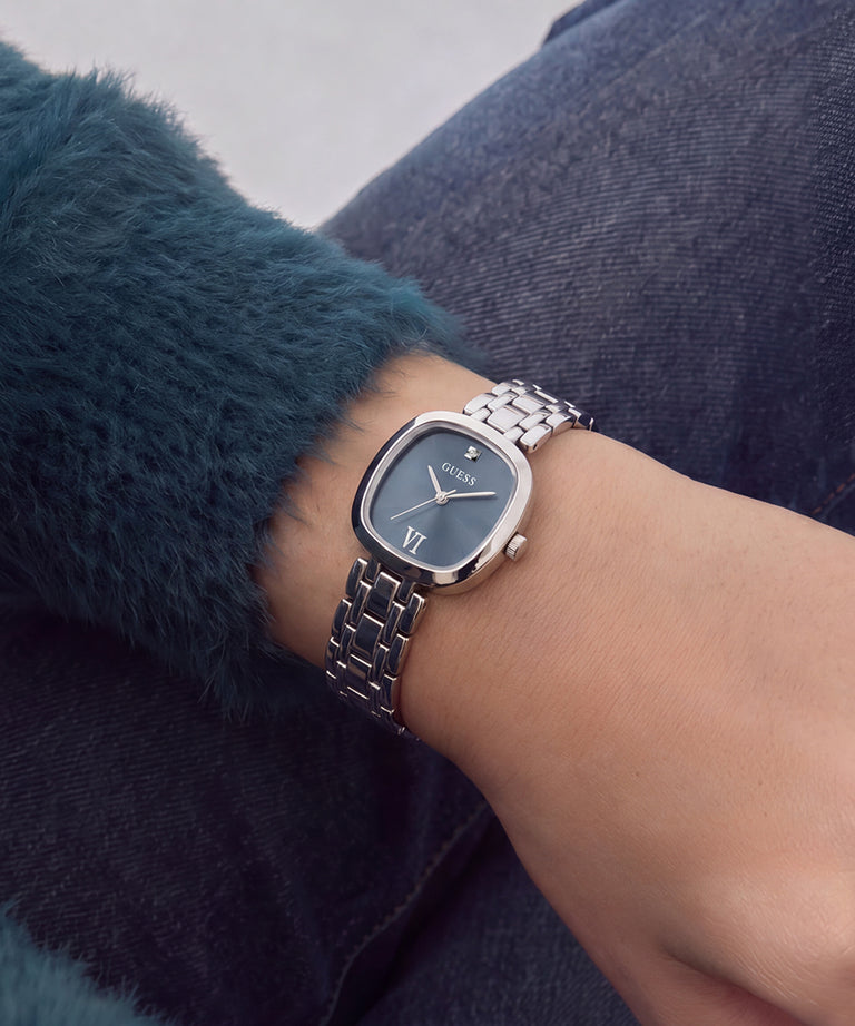 A silver-tone metal link watch with a dark blue dial and Roman numeral markers, worn on a person's wrist. The person is wearing a dark teal fuzzy sweater and dark blue denim jeans.