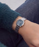 A silver-tone metal link watch with a dark blue dial and Roman numeral markers, worn on a person's wrist. The person is wearing a dark teal fuzzy sweater and dark blue denim jeans.