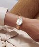 A close-up of a person's wrist wearing a gold-tone GUESS watch with a white dial and matching metal link bracelet, styled with a cream shirt and brown suede pants.