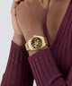 A gold-tone skeleton watch with a black dial and crystal-encrusted bezel, shown worn on a person's wrist. The person is wearing a ribbed burgundy long-sleeve top against a light gray studio background.