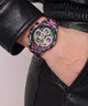 A close-up of a wrist wearing a colorful, highly detailed watch with a black leather jacket sleeve and black leather pants.