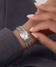A silver-tone rectangular wristwatch with a chunky chain-link bracelet, shown worn on a person's wrist with manicured hands resting nearby.