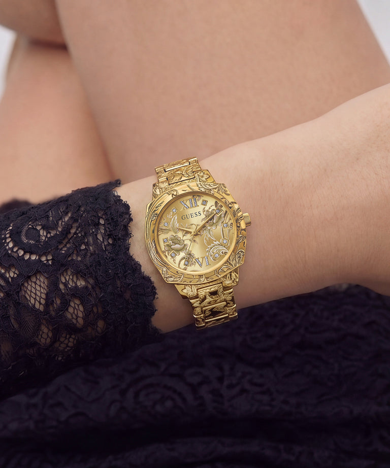 A gold-tone watch with a textured link bracelet and a detailed dial, shown worn on a person's wrist. The watch features Roman numeral hour markers and crystal accents.