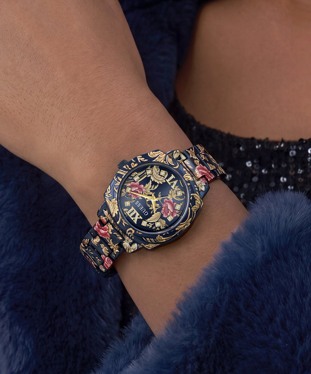 A close-up of a wrist wearing a decorative navy blue watch with gold floral and scroll motifs and pink rose accents, set against a dark blue furry garment.