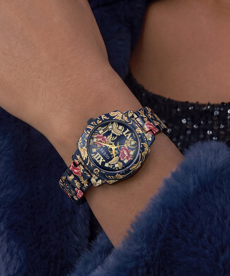 A close-up of a wrist wearing a decorative navy blue watch with gold floral and scroll motifs and pink rose accents, set against a dark blue furry garment.