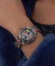 A close-up of a wrist wearing a decorative navy blue watch with gold floral and scroll motifs and pink rose accents, set against a dark blue furry garment.