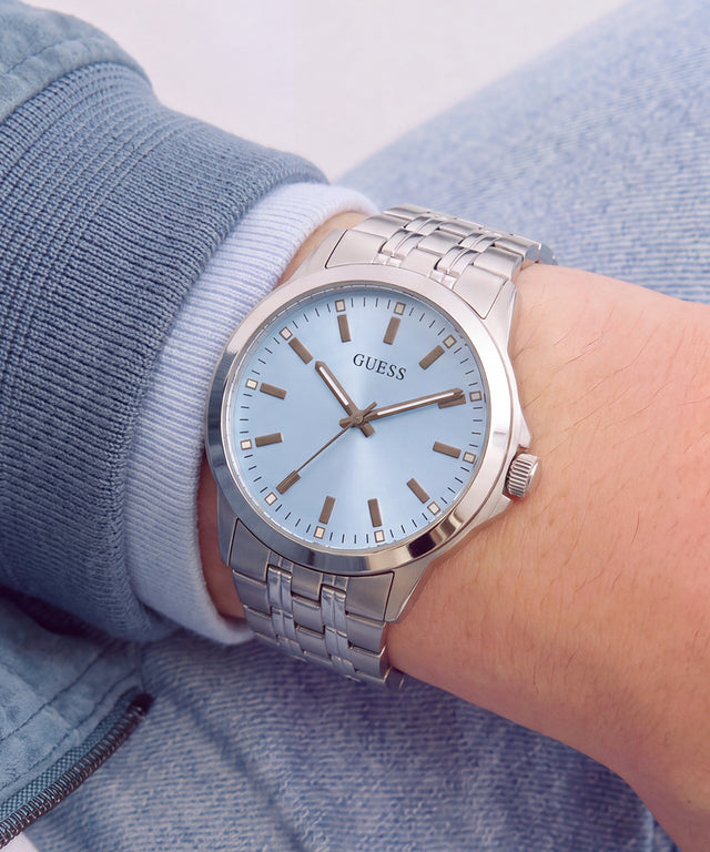 A wristwatch with a silver metal link bracelet and a light blue dial, worn on a person's wrist. The dial features the GUESS brand name and simple silver-tone hour markers and hands.