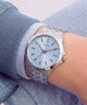 A wristwatch with a silver metal link bracelet and a light blue dial, worn on a person's wrist. The dial features the GUESS brand name and simple silver-tone hour markers and hands.