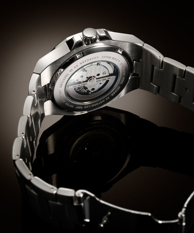 Close-up studio shot of the back of a stainless steel wristwatch with a transparent case back showing the mechanical movement, attached to a matching metal link bracelet on a dark reflective background.