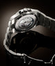 Close-up studio shot of the back of a stainless steel wristwatch with a transparent case back showing the mechanical movement, attached to a matching metal link bracelet on a dark reflective background.