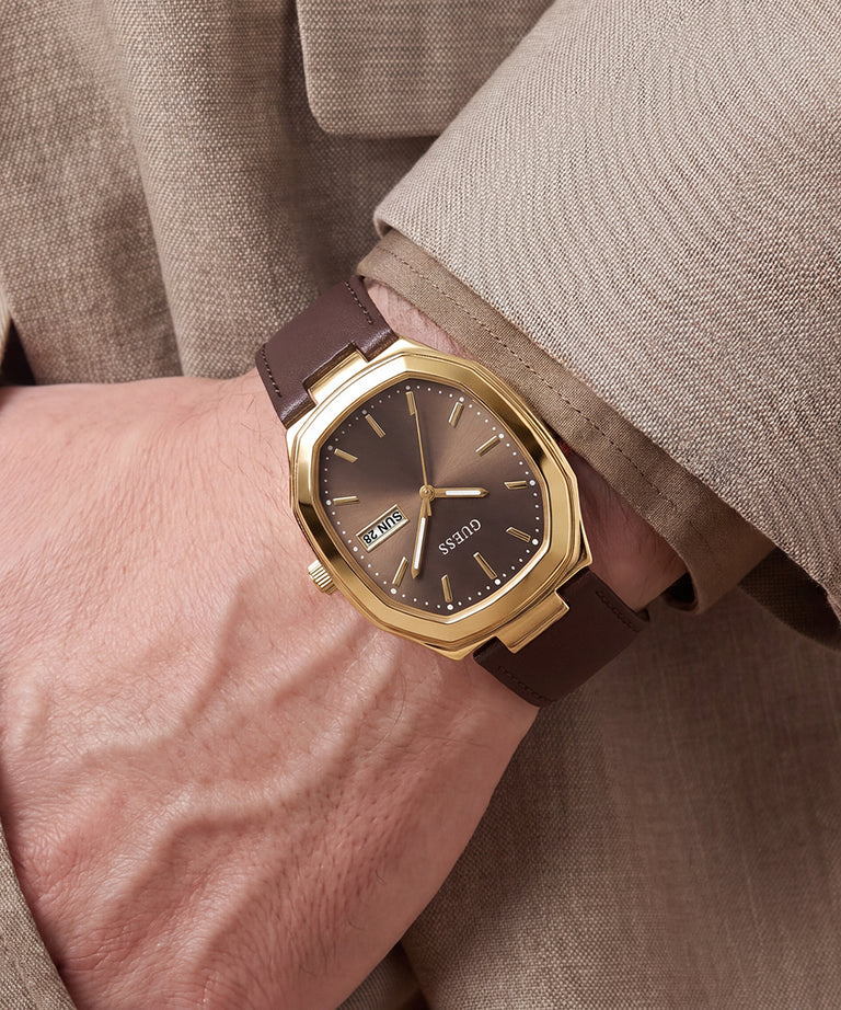 A close-up of a wrist wearing a Skoold watch with a gold-toned case, dark brown dial, and brown leather strap, set against a beige textured blazer.