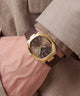 A close-up of a wrist wearing a Skoold watch with a gold-toned case, dark brown dial, and brown leather strap, set against a beige textured blazer.