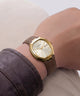 A wristwatch with a gold-tone octagonal bezel, cream dial, and brown leather strap, shown worn on a person's wrist.