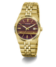 A gold-tone stainless steel wrist watch by Guess, featuring a fluted bezel, a gold-tone link bracelet, and a dark burgundy dial with gold-tone hour markers and hands.
