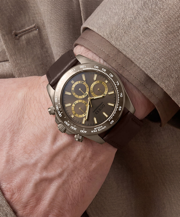 A close-up of a wrist wearing a guess chronograph watch with a dark brown leather strap, a brushed steel case, and a dark dial with gold subdials.