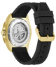 A close-up of a wristwatch showing the back case and strap. The watch has a gold-tone case with a transparent back revealing the mechanical movement, paired with a black textured silicone strap and a gold-tone buckle.