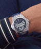A wristwatch with a silver-tone octagonal case, blue strap, and dark blue dial with multiple subdials, shown worn on a person's wrist. The dial shows the GUESS brand name.