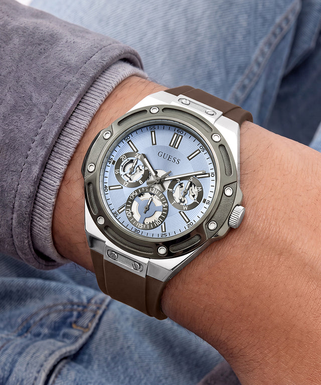 A close-up of a GUESS wristwatch with a brown rubber strap, silver-toned case, and light blue dial, worn on a person's wrist.