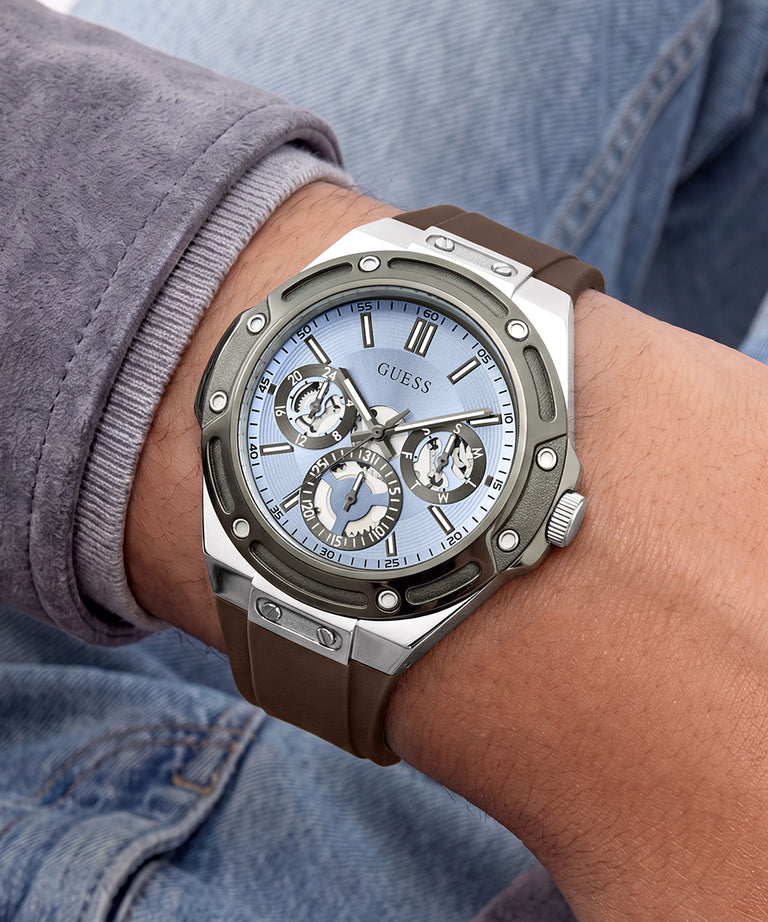 A close-up of a GUESS wristwatch with a brown rubber strap, silver-toned case, and light blue dial, worn on a person's wrist.
