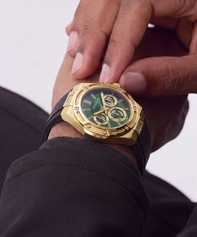 A close-up of a wrist wearing a gold-tone Hublot watch with a green dial and black strap, with fingers adjusting the crown.