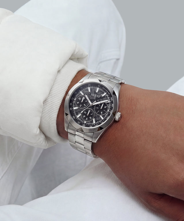 A silver stainless steel wristwatch with a black dial and integrated metal bracelet, shown worn on a person's wrist. The person is wearing a white long-sleeve shirt, and the background is a soft, neutral gray.