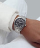 A silver stainless steel wristwatch with a black dial and integrated metal bracelet, shown worn on a person's wrist. The person is wearing a white long-sleeve shirt, and the background is a soft, neutral gray.