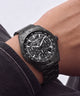 A close-up of a black dial steel chronograph watch with a matching black metal link bracelet, worn on a person's wrist.