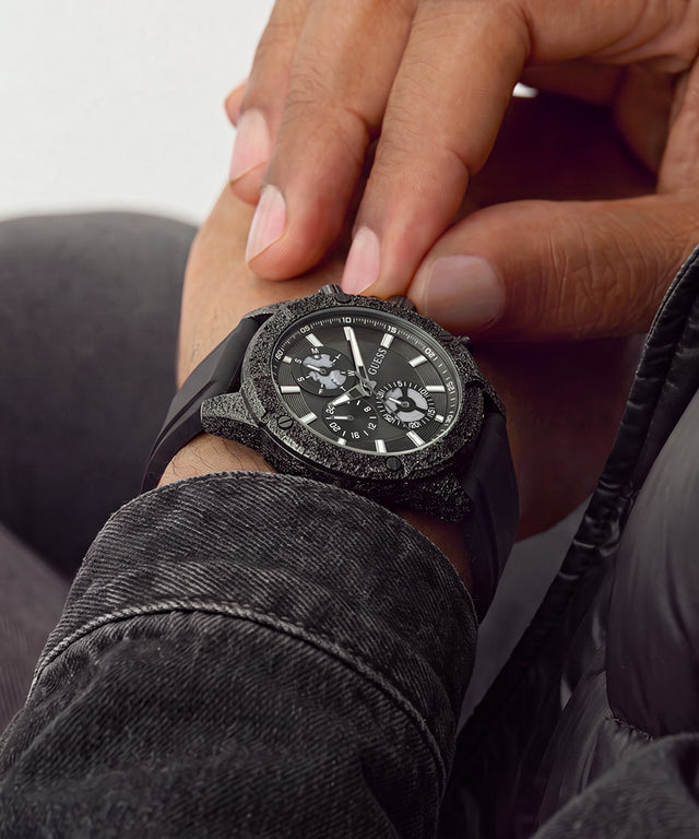 A close-up lifestyle shot of a black chronograph watch with a textured, stone-like bezel and a black rubber strap, worn on a person's wrist with their hand resting near it.